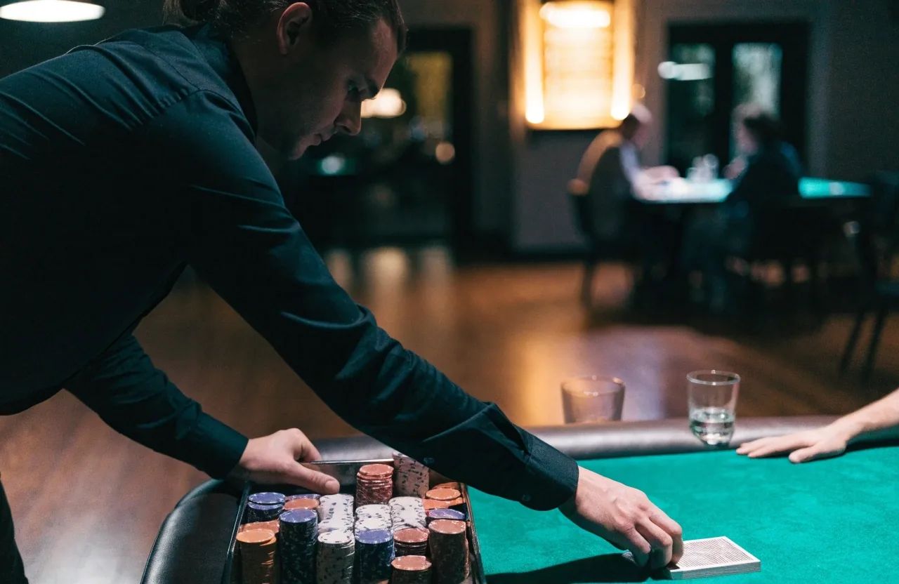 Pokerový stůl v herně večer — dealer rozdává karty, na railu stojí sloupce žetonů